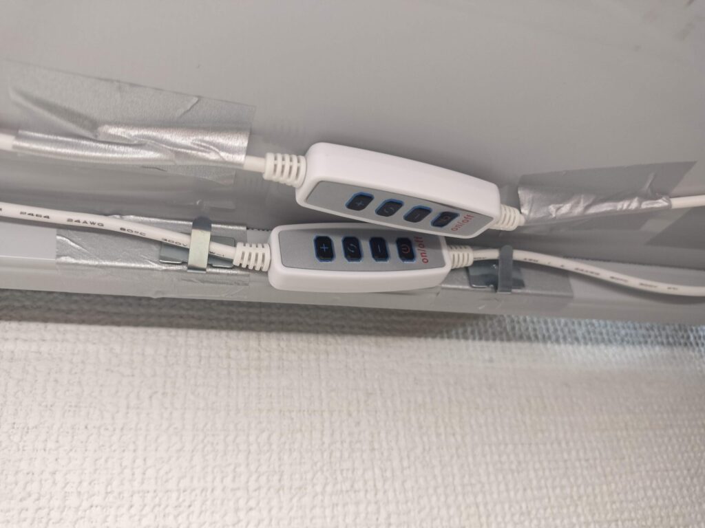 絵本棚の棚板裏面にコードフックでLEDライトのコードとコントローラーをまとめた様子、手前のコントローラーはテープのみで固定。